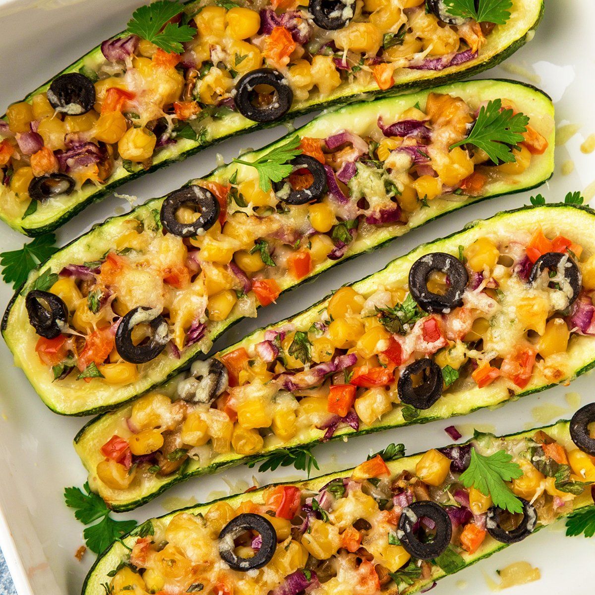 Stuffed zucchini boats filled with corn, red pepper, cheese, and olives, in a white baking dish.