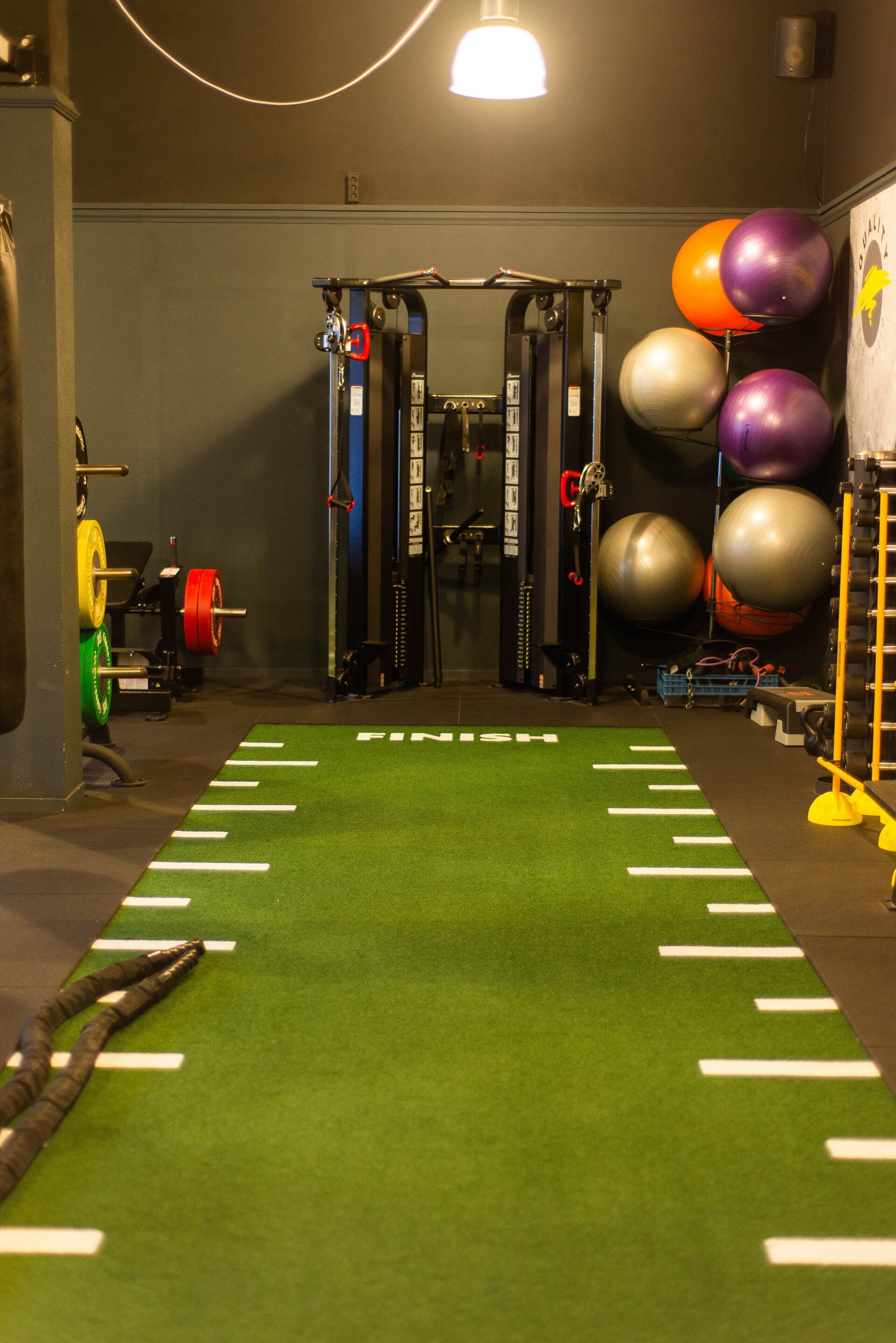 Sportschoolinterieur met groene kunstgrasbaan, fitnessapparatuur en medicijnballen.