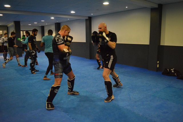 Twee mannen oefenen kickboksen in een sportschool, met beschermende kleding aan. Blauwe vloer, andere mensen op de achtergrond.