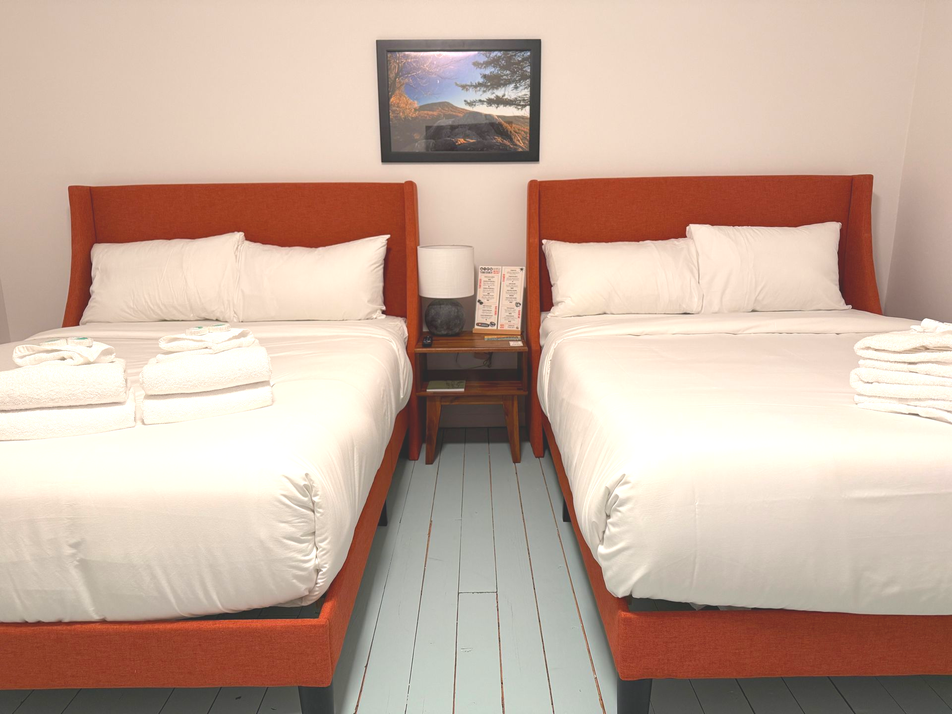 Two orange-framed double beds with white linens in a room with a painting, nightstand, and light blue floor.