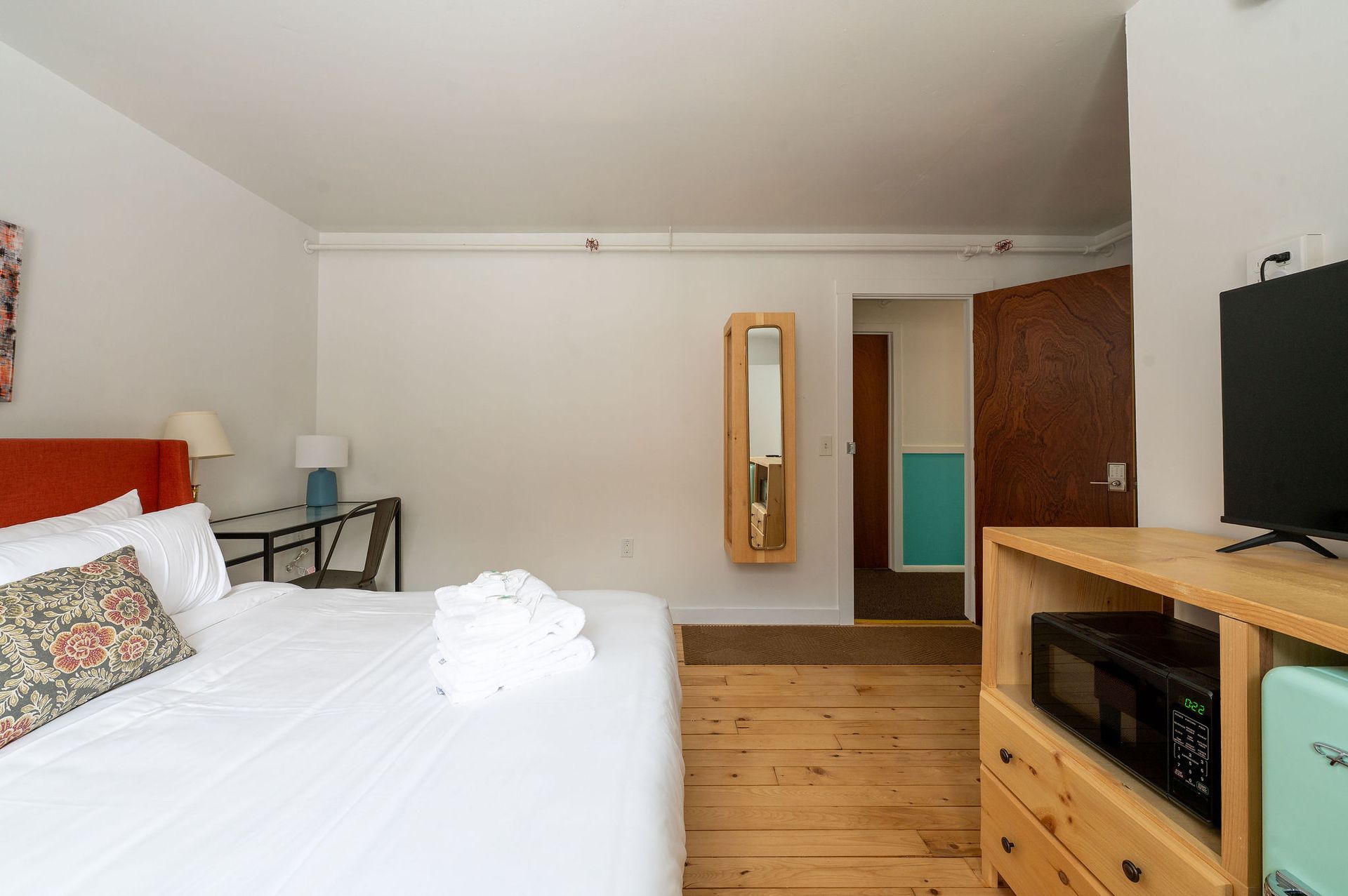 Bedroom with white bedding, wooden floors, a small TV, and a microwave. Doorway leads to a bathroom.