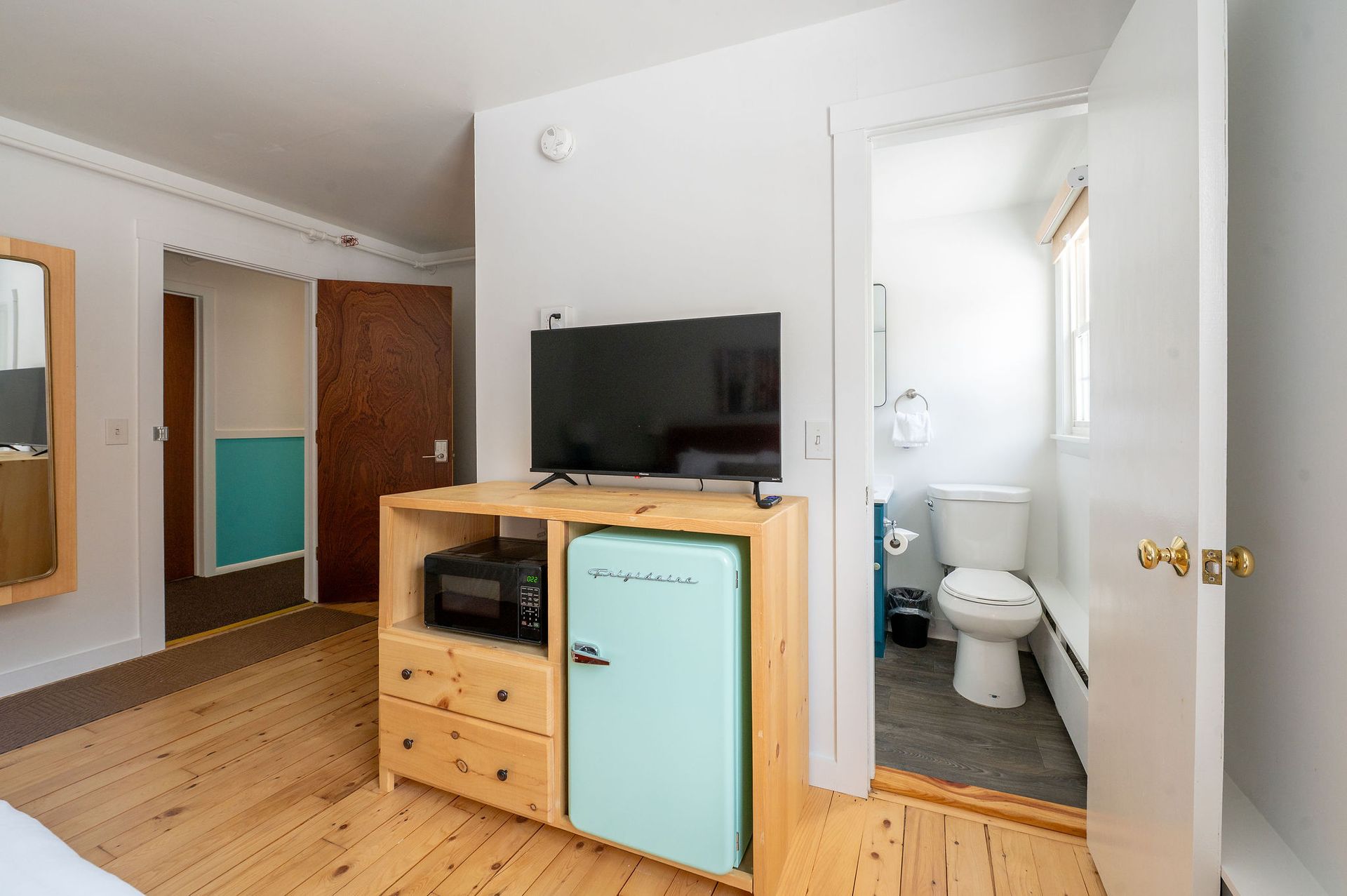 Bedroom with light wood floor, TV, and turquoise mini-fridge built into a wooden cabinet. The bathroom is visible.