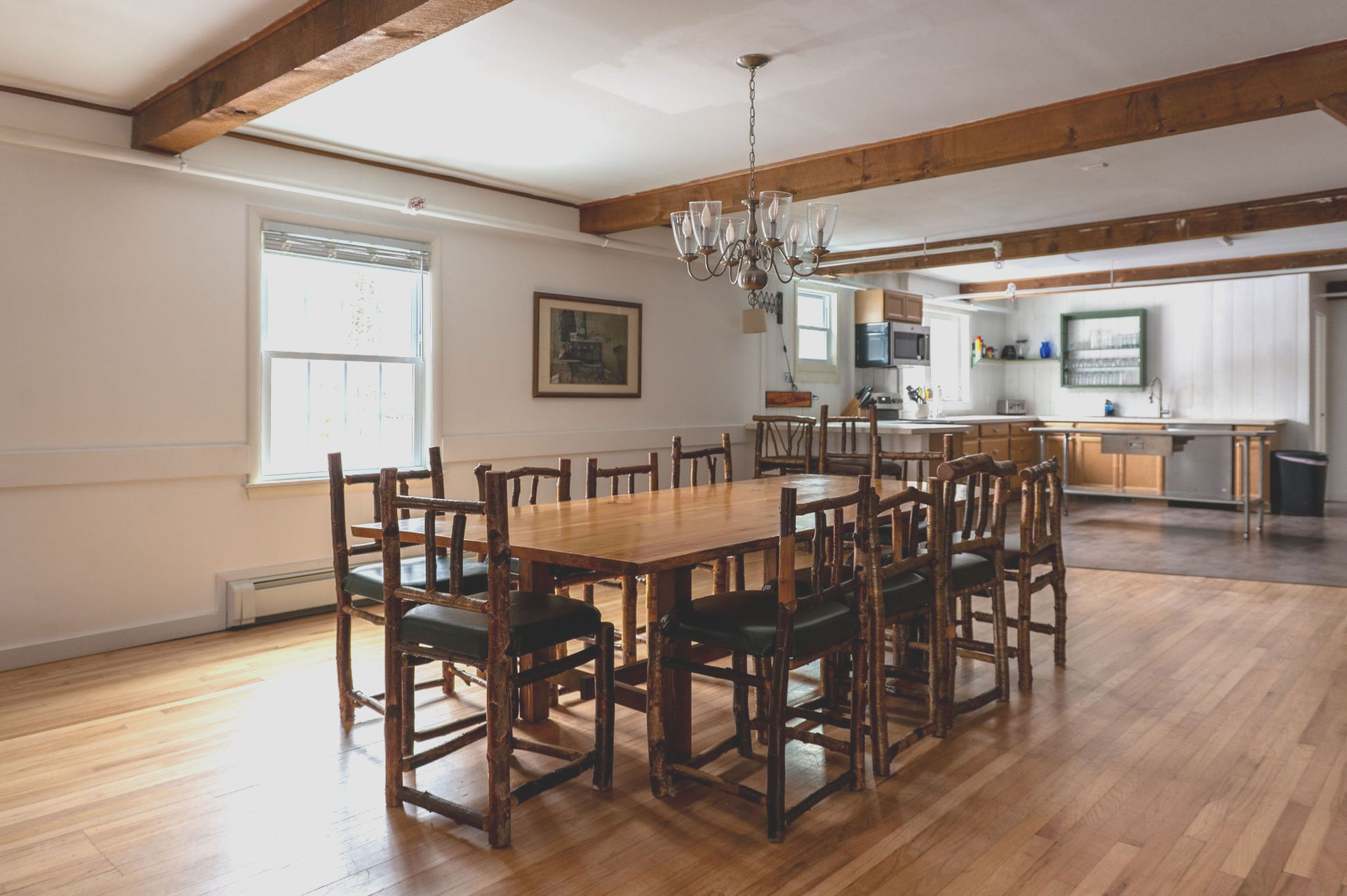 Large dining room with long wooden table, chairs, and chandelier. Kitchen visible in the background.