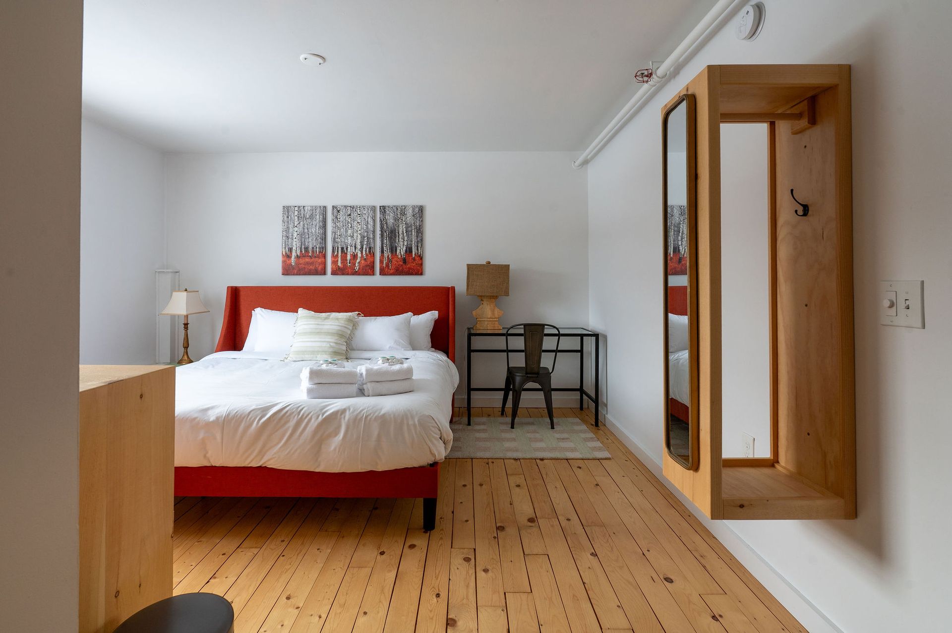 Bedroom with red bedframe, artwork, desk, wooden floor, and a mirror on the wall.