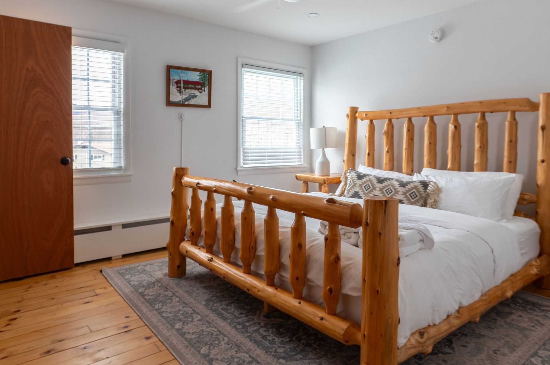 Bedroom with a wooden log bed, two windows, a painting, and a wooden door.