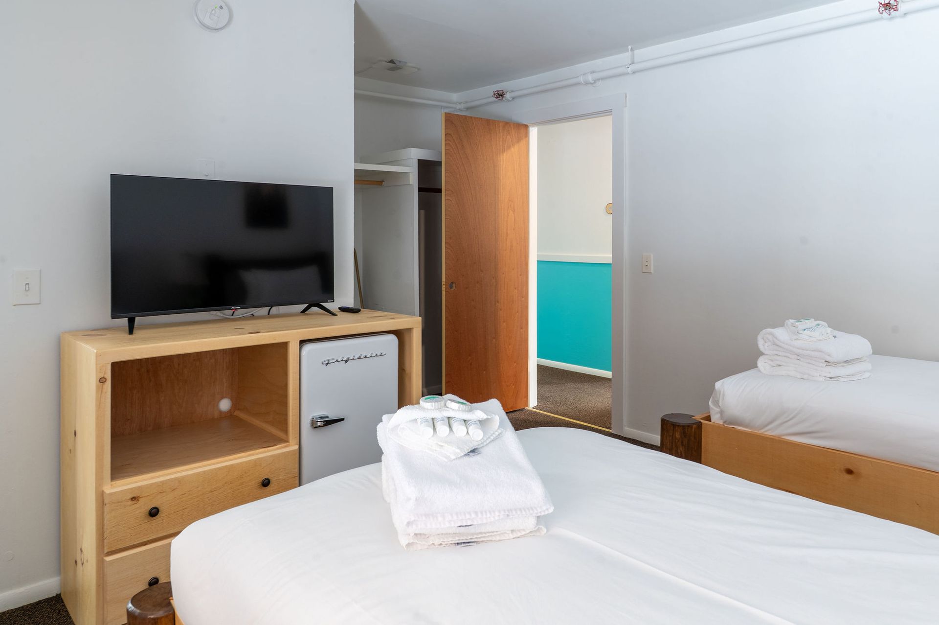 Hotel room with two beds, TV, and mini-fridge on a wooden stand. Doorway to another room.