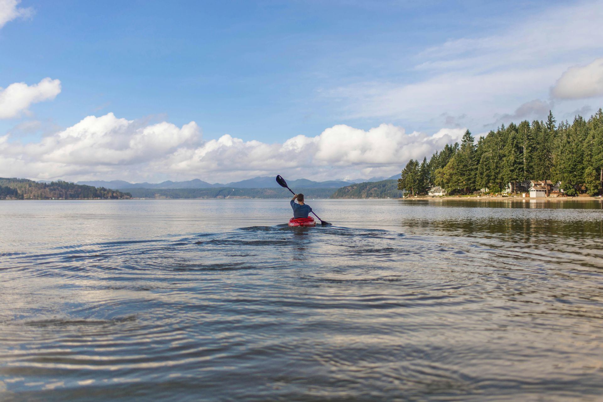 Kayaker on orange kayak, paddles on calm lake, mountain backdrop.