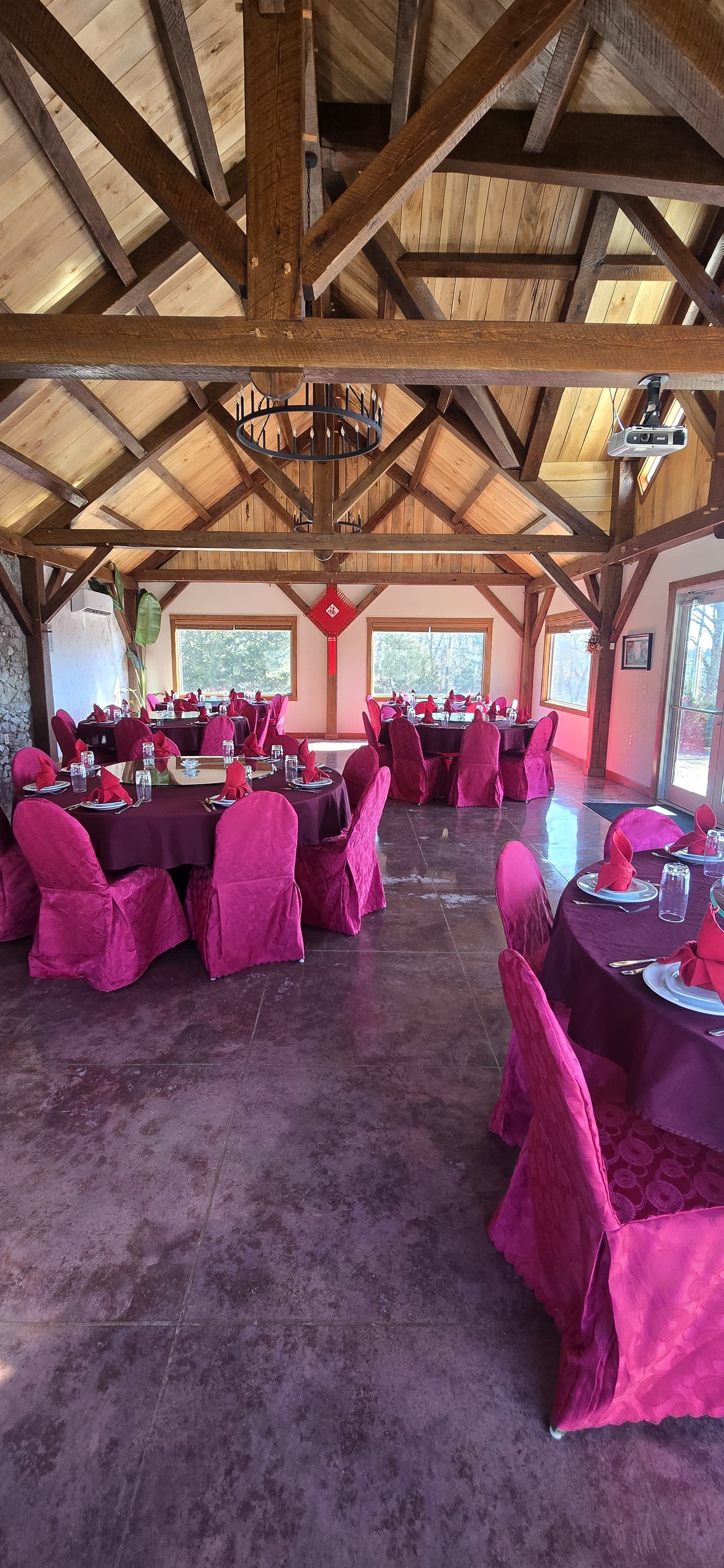Image of spacious room with a peaked ceiling and wooden beams, three round tables, and pink/red chairs.