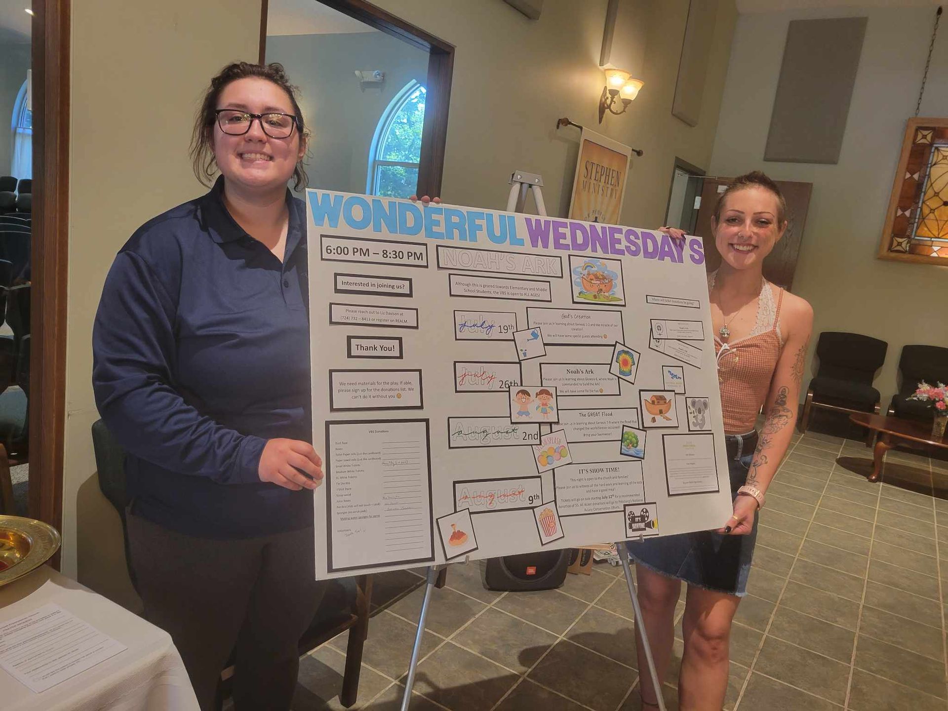 Two women holding a colorful “Wonderful Wednesday” poster in a church hall