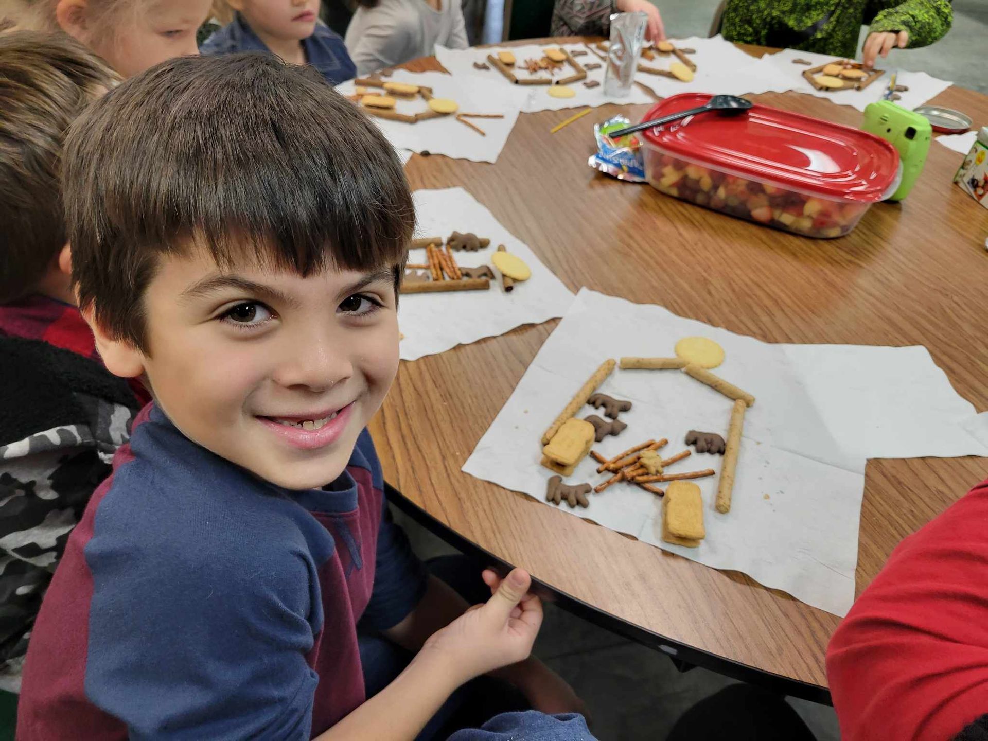 Child smiling at a classroom table with craft materials and papers scattered around.