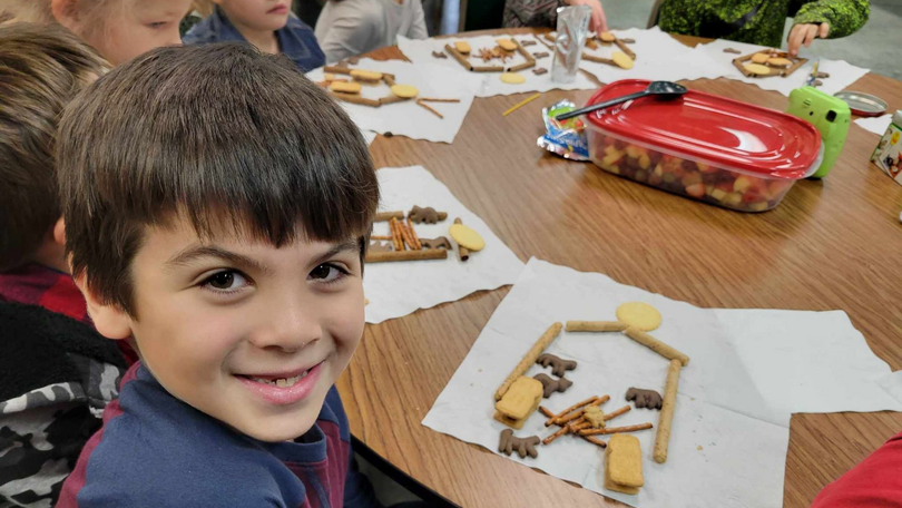 Smiling child at a classroom table with paper and snack pieces, other children and a red tray nearby.