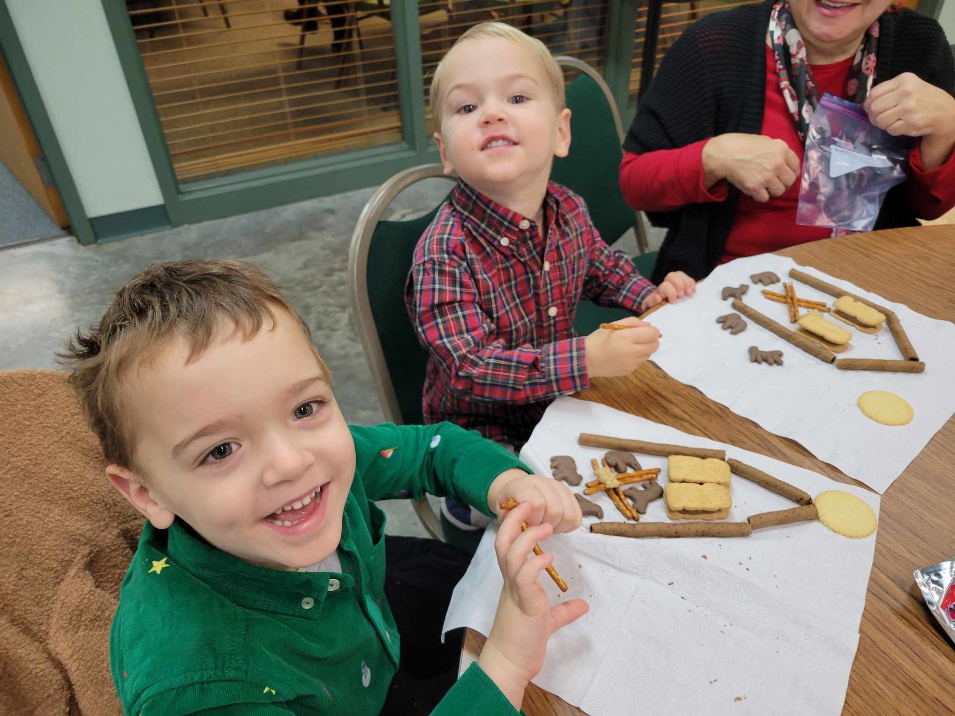 Children decorating gingerbread cookie houses at a table with icing and candy, smiling at the camera