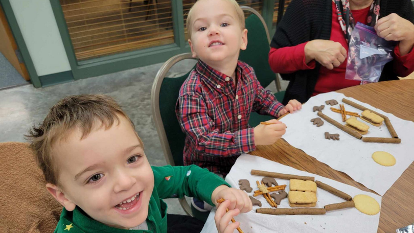Children decorating gingerbread houses at a table, with one smiling at the camera.
