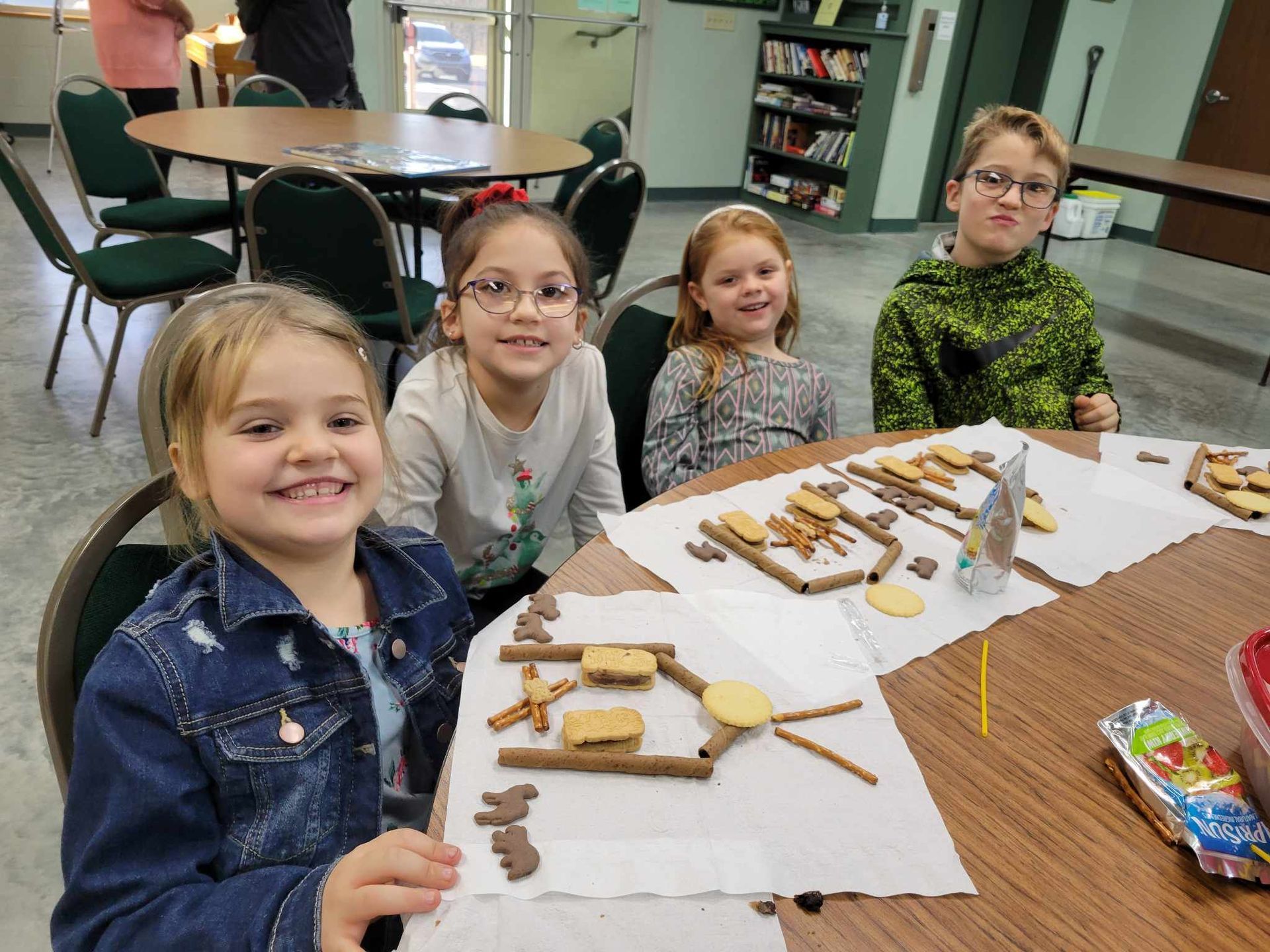 Children seated at a table, smiling while making craft projects with sticks and paper.