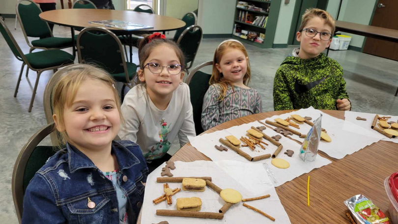 Four children smiling at a table with craft materials and unfinished popsicle-stick projects