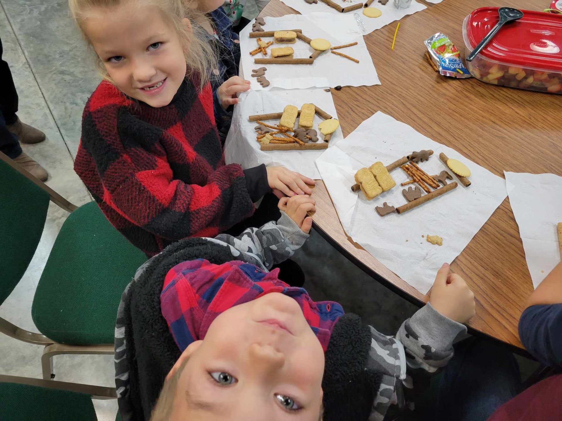 Two children at a table decorating gingerbread houses with candy and icing, smiling at the camera