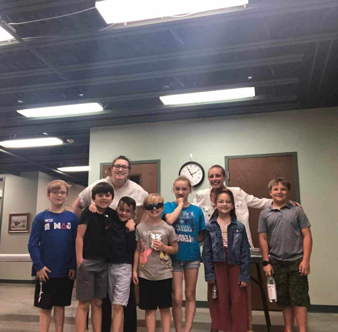 Group of eight people standing indoors, smiling in front of a wall clock and doors.