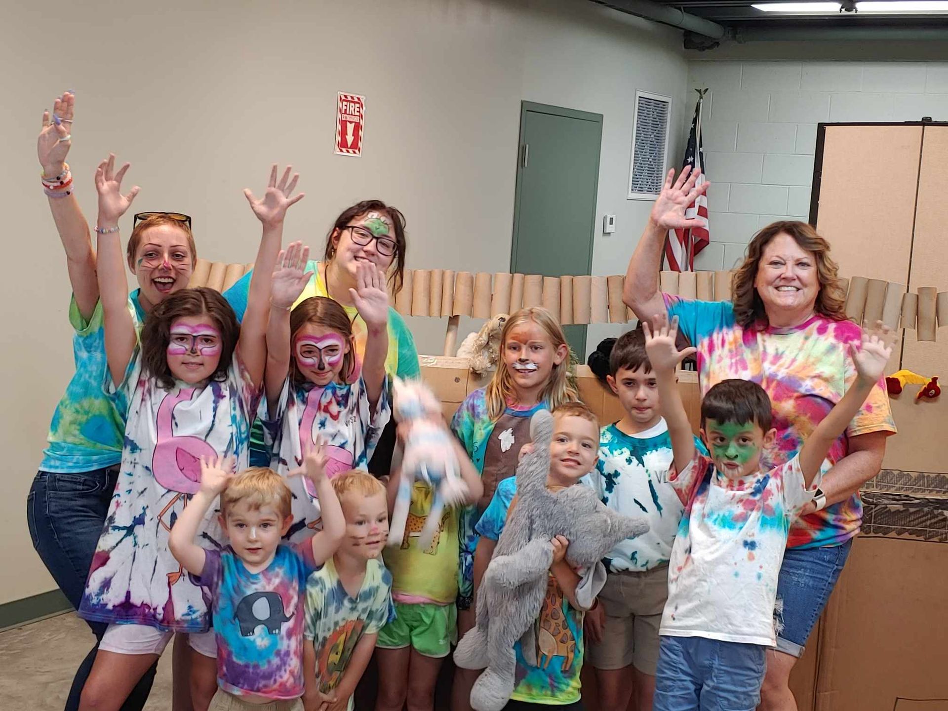 Group of smiling people in tie-dye shirts, raising their hands amid colorful powder indoors