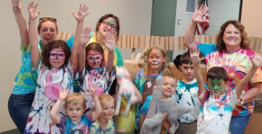A group of people in tie-dye shirts smile and wave with colorful paint-splattered hands outdoors.