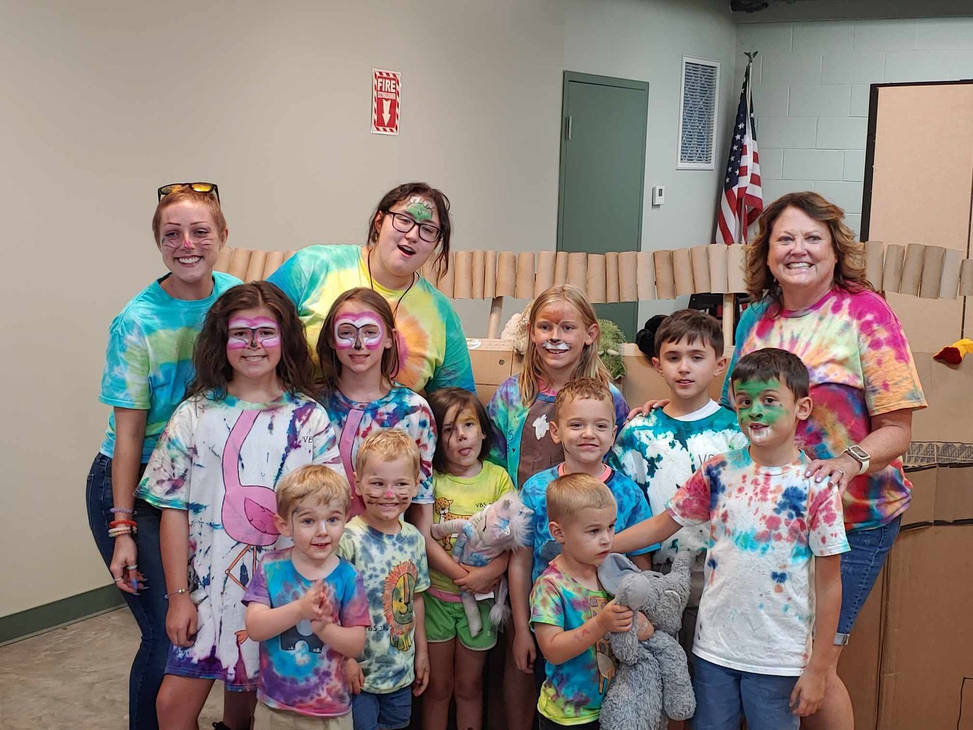 Group of people in tie-dye shirts posing indoors, smiling in a room with a U.S. flag.