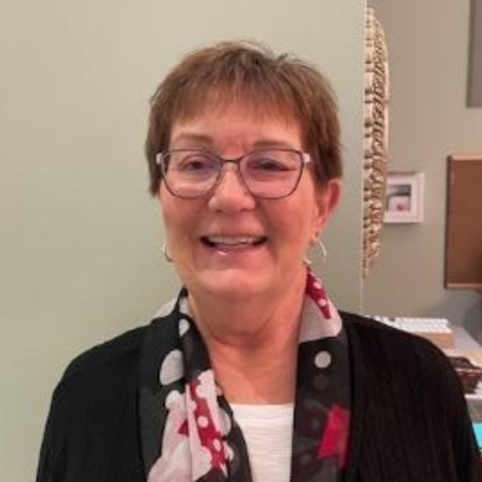 Smiling person wearing glasses, black cardigan, and patterned scarf in an indoor setting