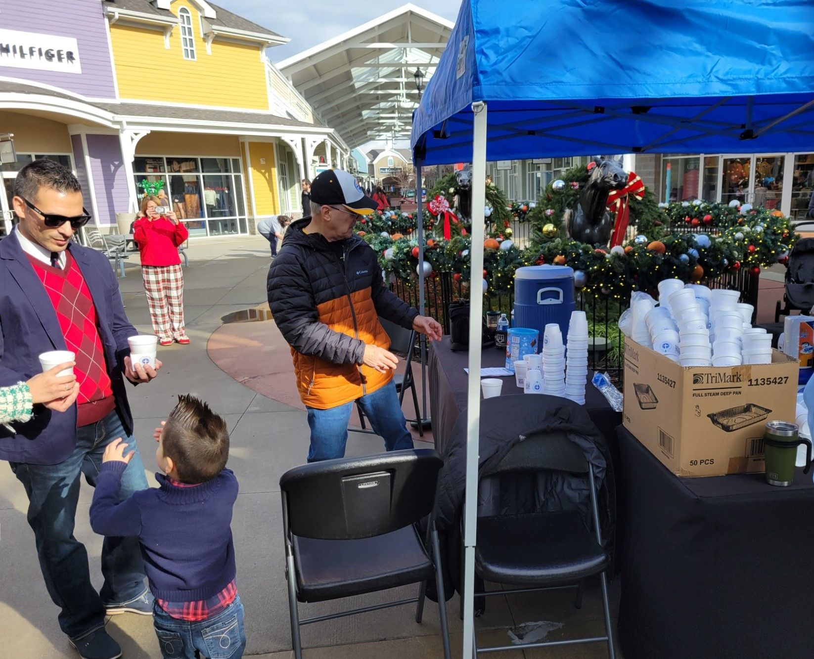 People at an outdoor market booth with blue canopy, tables of cups, and a child reaching toward an adult