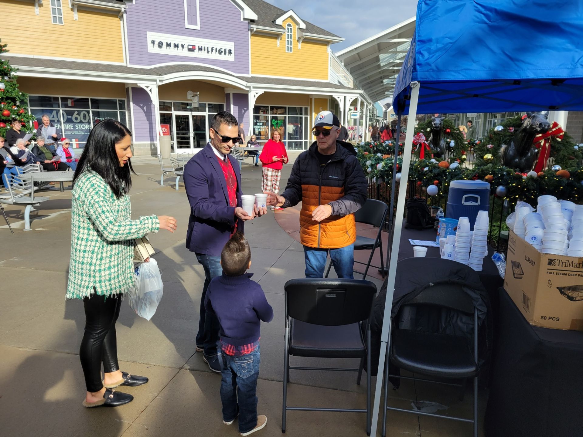 People chatting beside a blue canopy at an outdoor holiday market, with a child in the foreground.
