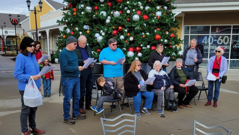 People gathered outdoors by a decorated Christmas tree, some standing and some seated, near a building.