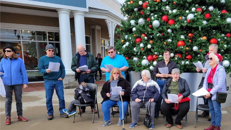 People gathered outdoors by a large decorated Christmas tree, some seated and some standing, holding papers.