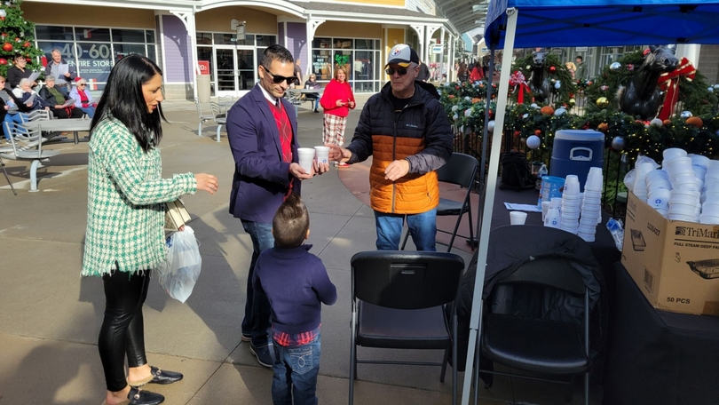 People chatting outdoors near a decorated cart, with a child facing them in a sunny plaza.