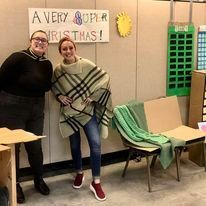 Two people posing beside a “A VERY SUPER BRATMAS!” sign, with chairs and green paper decorations.