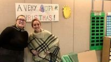 Two people smiling in a room beside a “A Very Super Christmas!” sign and green storage bins.