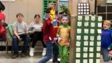 Children in colorful costumes perform indoors beside seated adults and a green checkerboard prop