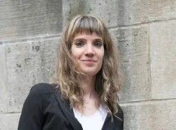 Woman standing against a concrete wall, wearing a black blazer and white top, with a slight smile.