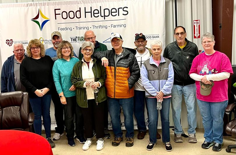 Group of people posing in front of a Food Helpers banner, holding bags and smiling indoors.