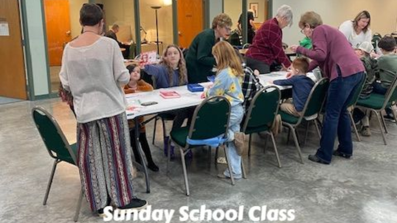 Sunday school class gathered around tables in a bright room, with adults and children working together.