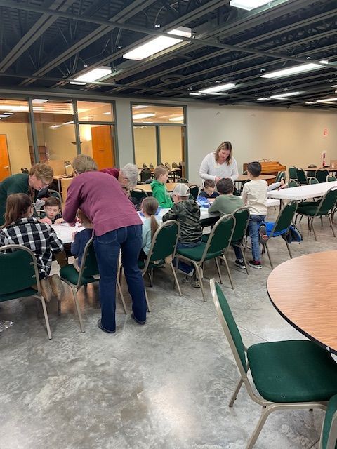 People gathered around tables in a community hall, chatting and working together under bright overhead lights.
