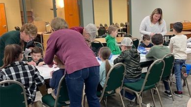 People gathered around tables in a classroom, working together on an activity with a teacher assisting.