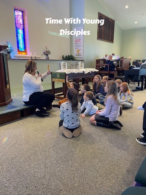 Children sit on carpet listening to a speaker in a church room with a projected “Time With Young Disciples” slide.