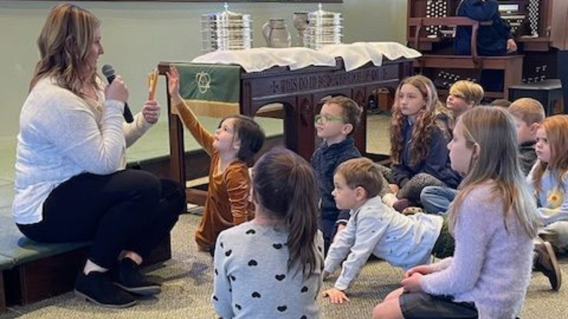 Teacher showing a child a small object while a group of children sit on the floor in a classroom