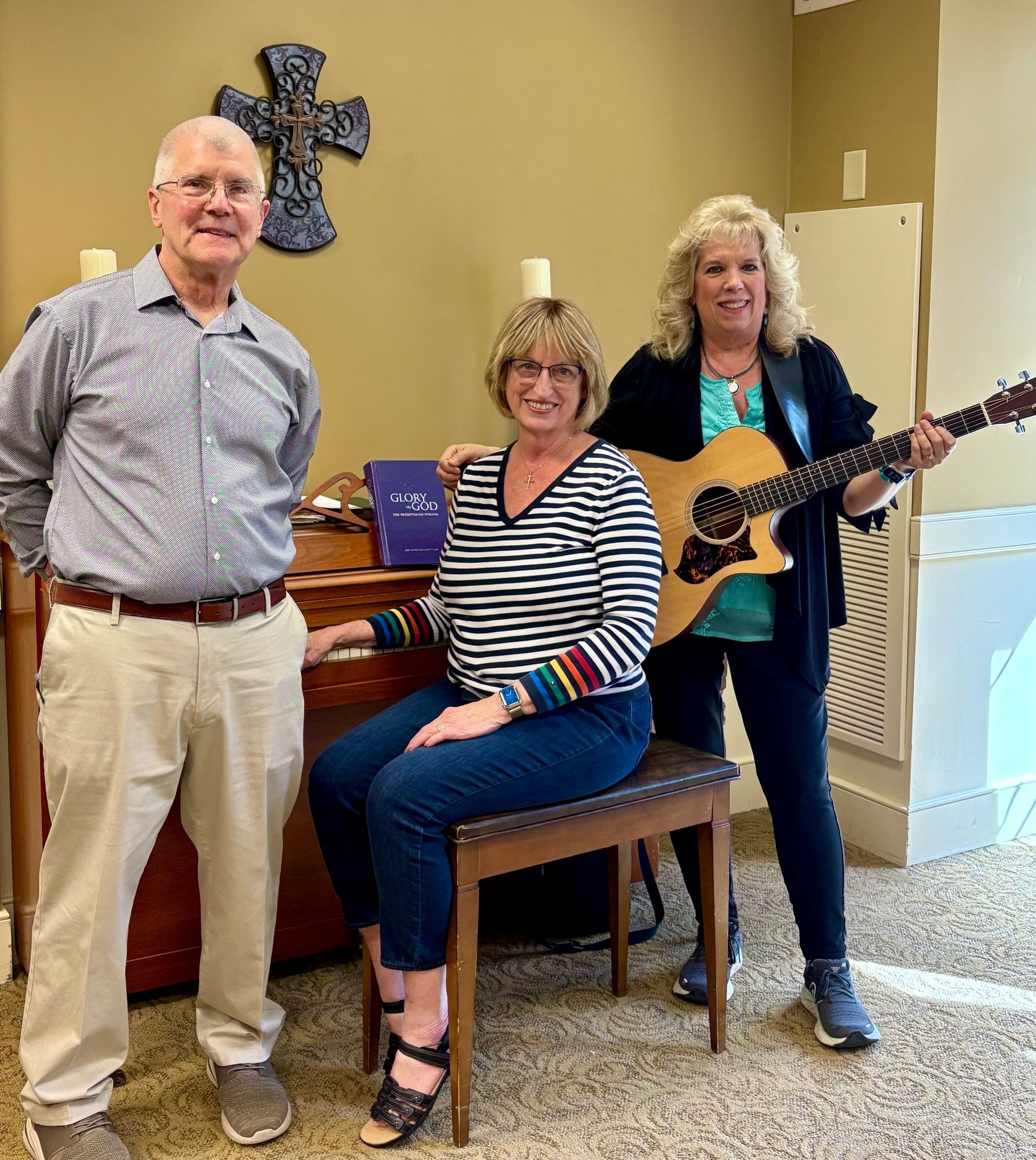Three adults posing indoors; one holds a guitar, standing beside a seated woman and a man.