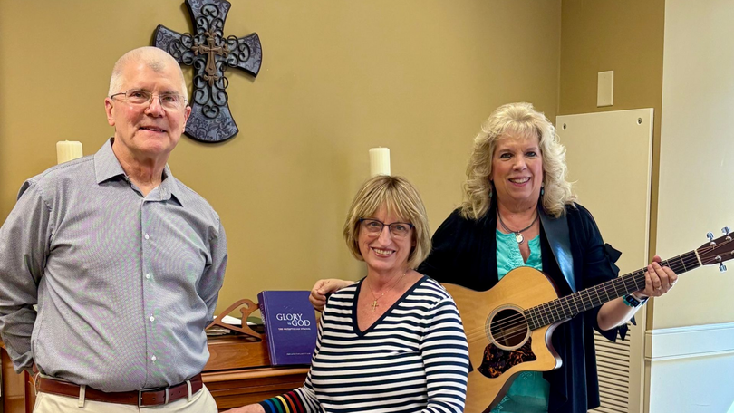 Three people stand in a church-like room, with one holding a guitar and a cross on the wall behind them.