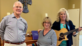 Three people smiling indoors, one holding a guitar, in a room with a piano and candles.