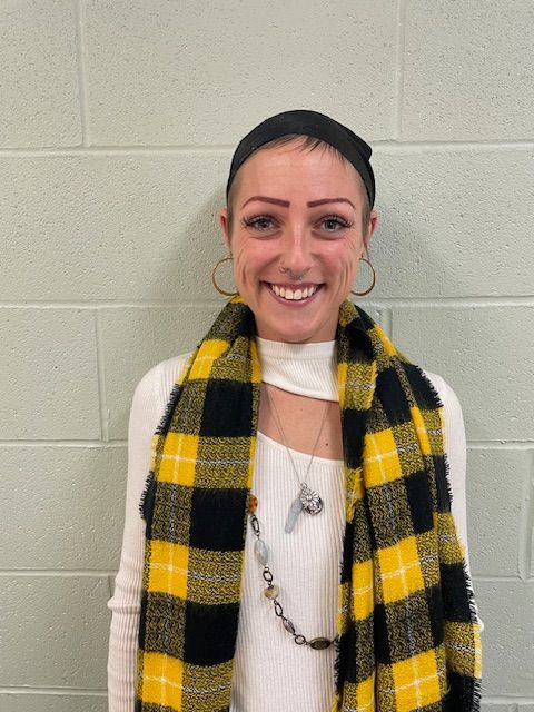 Smiling person in a black-and-yellow plaid scarf standing against a light gray brick wall