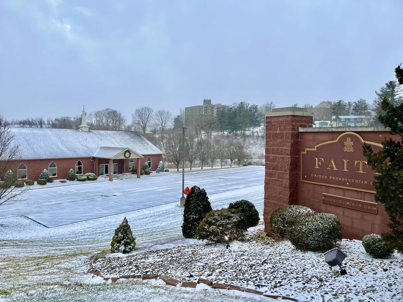 Snowy campus entrance with brick sign, landscaped shrubs, and a person walking near a building under an overcast sky