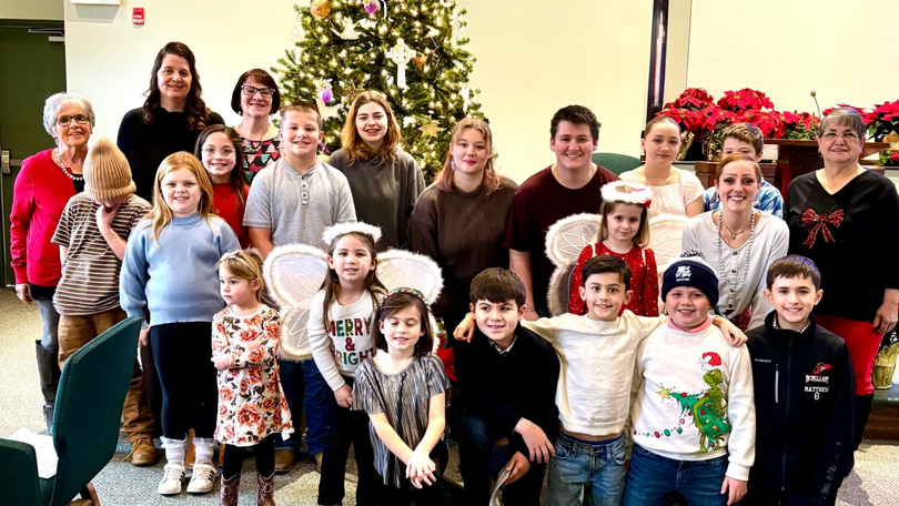 Group of children and adults posing indoors beside Christmas trees and holiday decorations.