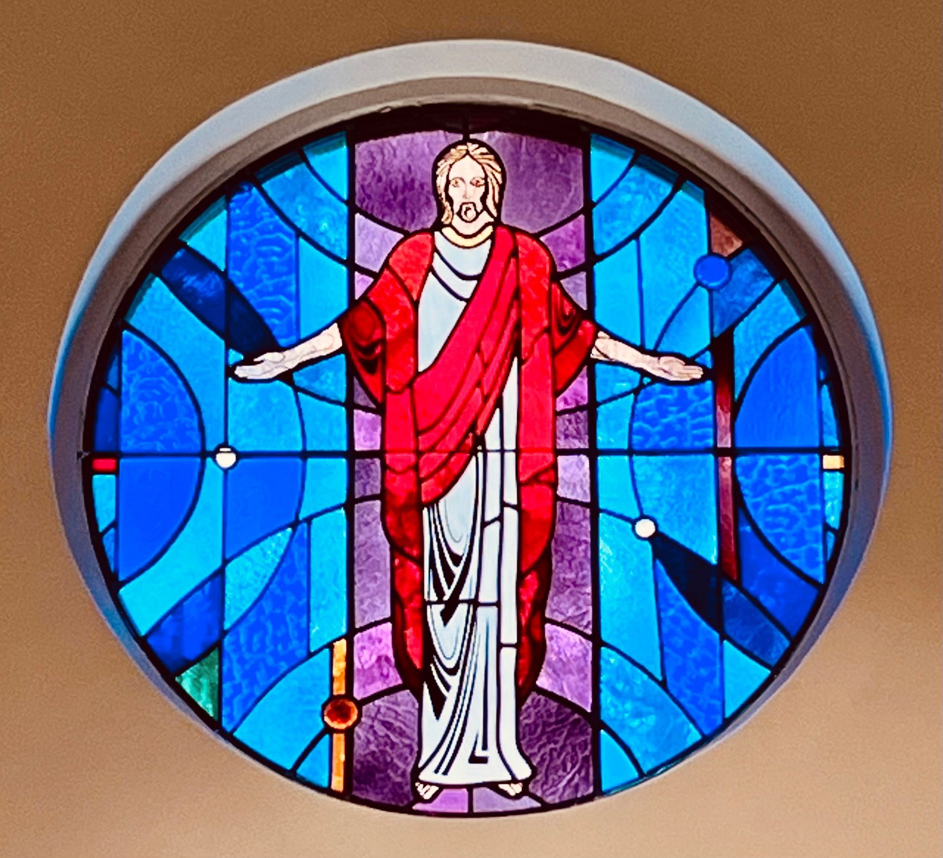 Stained glass window of Jesus in a red robe with outstretched arms, surrounded by blue and purple panels