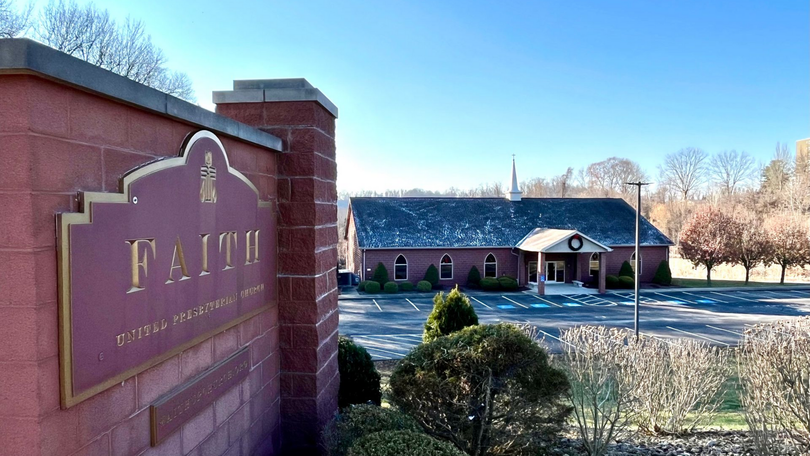 Brick entrance sign for Faith with a building and parking lot in the background under a clear sky