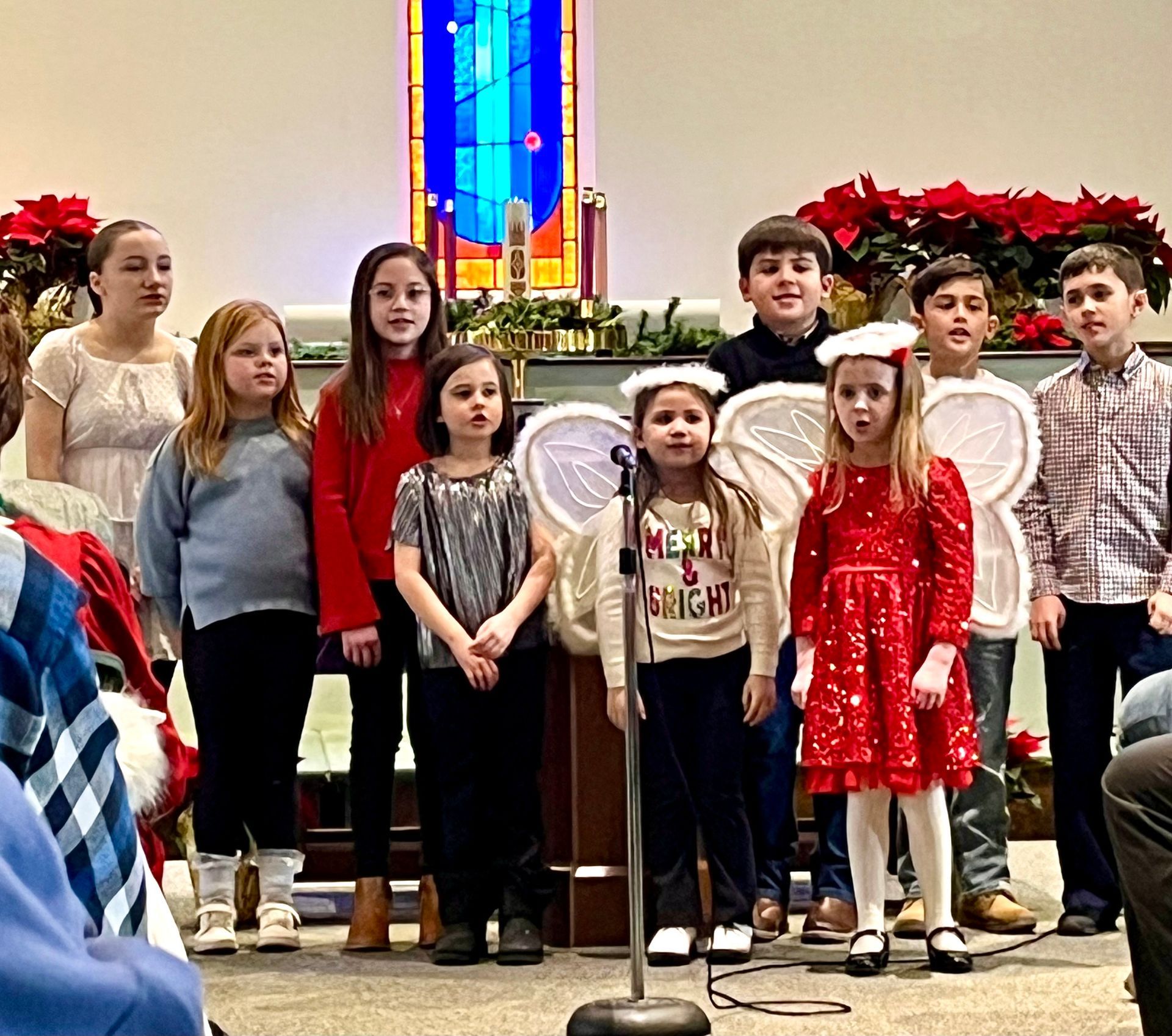 Children standing in a church near a pulpit, with Christmas decorations and stained glass behind them
