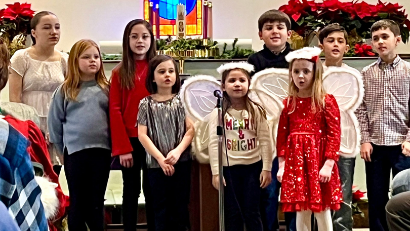 A group of children stand on a stage in colorful holiday outfits, posing in front of festive decorations.